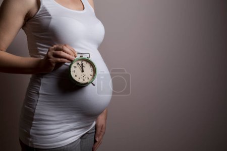 Téléchargez les photos : Gros plan du ventre d'une femme enceinte, debout à côté d'un fond gris et tenant une horloge représentant une proximité d'une date d'échéance - en image libre de droit