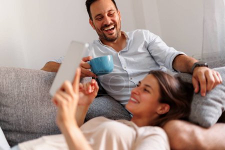 Photo for Beautiful young couple in love relaxing at home, surfing the Internet using tablet computer and drinking coffee in the morning - Royalty Free Image