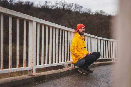 Téléchargez les photos : Jeune homme actif accroupi et penché à la clôture du pont tout en prenant une pause de jogging, faire sa séance d'entraînement matinale - en image libre de droit