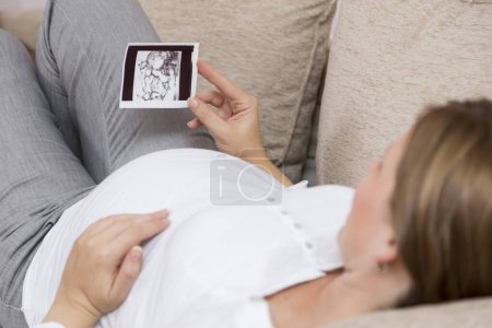 Téléchargez les photos : Femme enceinte allongée sur un canapé dans un salon de son appartement, tenant et regardant la photo échographique de son bébé - en image libre de droit