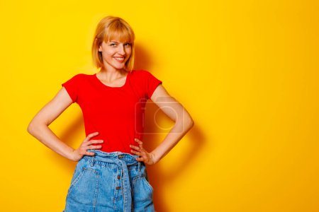 Photo for Beautiful young blond woman wearing trendy summer wear holding hands on hips on yellow colored background with copy space - Royalty Free Image
