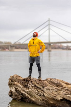 Téléchargez les photos : Homme athlétique debout sur un tronc d'arbre tombé, se détendre et faire une pause tout en faisant du jogging au bord de la rivière par une journée d'automne couvert - en image libre de droit