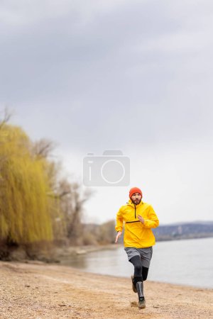 Foto de Hombre activo trotando junto al río en un día nublado de otoño lluvioso - Imagen libre de derechos