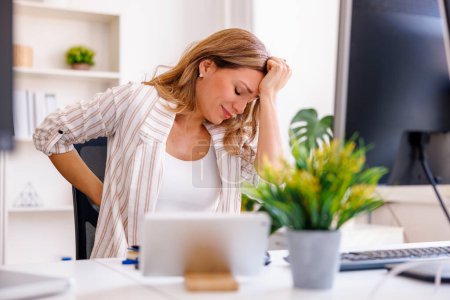 Téléchargez les photos : Femme ayant mal à la tête et mal au dos alors qu'elle était assise à son bureau, fatiguée après une longue journée de travail - en image libre de droit