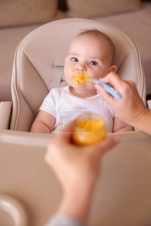 Photo for High angle view of mother feeding cheerful baby boy with porridge, baby sitting in high chair all messy and staied smiling and eating - Royalty Free Image