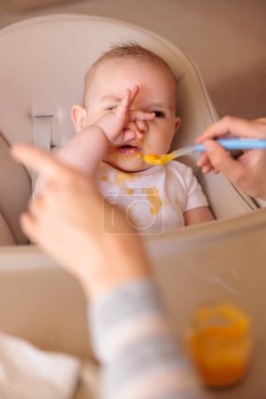 Photo for Little baby boy sitting in high chair making funny faces and refusing to eat pumpkin porridge while mother trying to feed him using spoon - Royalty Free Image