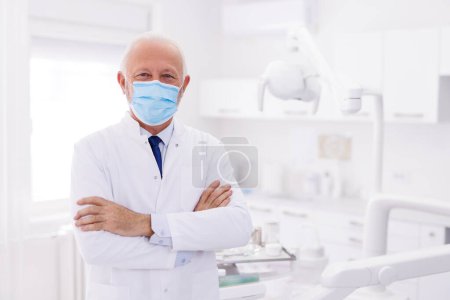 Téléchargez les photos : Portrait d'un médecin homme senior portant un masque protecteur médical debout à l'hôpital avec les bras croisés - en image libre de droit