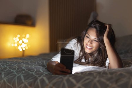 Téléchargez les photos : Joyeux jeune femme couchée dans le lit la nuit, surfer sur le net à l'aide d'un téléphone intelligent - en image libre de droit