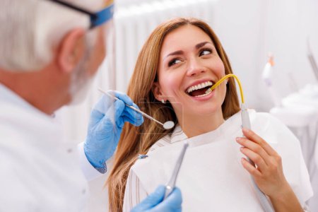 Dentista que fija el diente del paciente en la clínica dental usando taladro dental y espejo en ángulo, paciente sentado en la silla dental y sosteniendo el eyector de saliva para succión