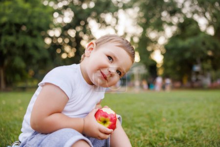 Photo for Portrait of cute little baby boy sitting on grass while on picnic with his parents eating an apple and smiling - Royalty Free Image