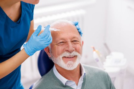 Foto de Médico aplicando rellenos dérmicos al paciente masculino mayor; hombre mayor recibiendo ácido hialurónico o tratamiento antiedad botox - Imagen libre de derechos