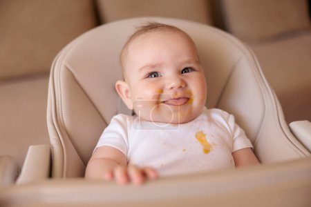 Cute little baby boy laughing and playing in high chair all messy and stained after eating porridge