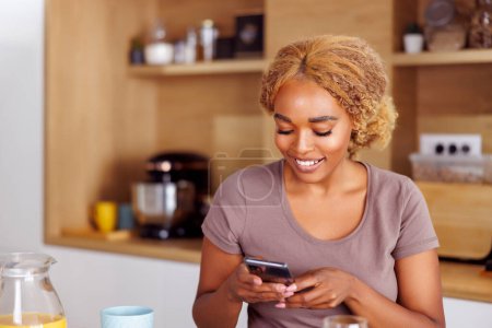 Téléchargez les photos : Belle jeune femme assise au comptoir de la cuisine prenant le petit déjeuner et surfant sur le net en utilisant un téléphone intelligent, se détendre à la maison le matin - en image libre de droit
