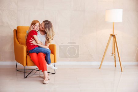 Téléchargez les photos : Mère et fille assises dans un fauteuil, mère tenant son bel enfant sur ses genoux et l'embrassant, appréciant passer du temps libre ensemble à la maison - en image libre de droit