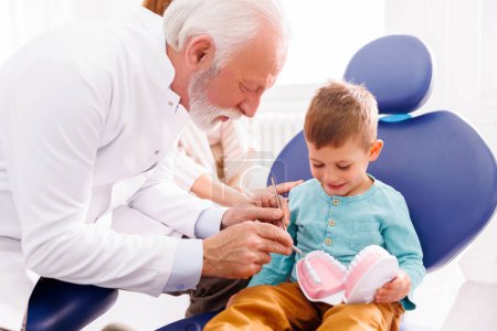 Foto de Lindo niño sentado en la silla dental, mientras que el dentista masculino senior demostrando procedimiento de chequeo en el modelo de mandíbula humana de plástico - Imagen libre de derechos
