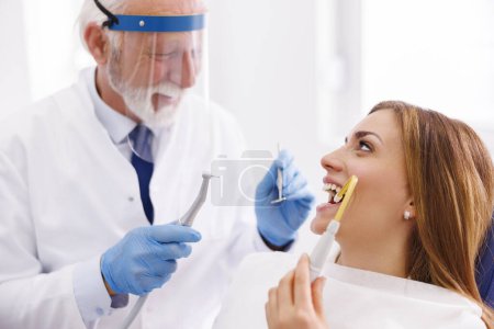 Foto de Dentista que fija el diente del paciente en la clínica dental usando taladro dental y espejo en ángulo, paciente sentado en la silla dental y sosteniendo el eyector de saliva para succión - Imagen libre de derechos