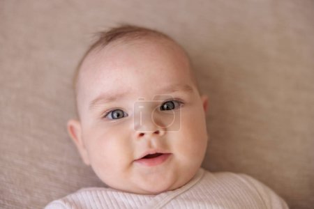 Téléchargez les photos : Vue du dessus portrait de mignon bébé garçon ludique avec eczéma éruption cutanée sur le front souriant et regardant vers la caméra tout en étant couché sur le canapé - en image libre de droit