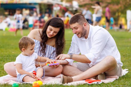 Photo for Beautiful young parents having picnic in the park with their cute little baby boy, sitting on the blanket and playing with colorful balls - Royalty Free Image