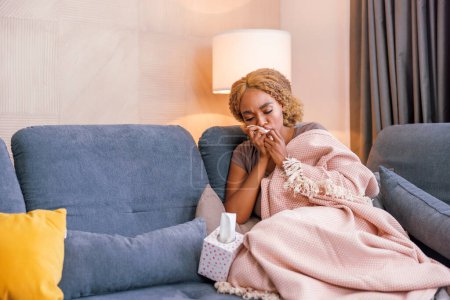 Foto de Mujer acostada enferma en casa cubierta con manta en el sofá de la sala de estar, sufriendo de dolor con fiebre y gripe, tos, estornudos y sonarse la nariz - Imagen libre de derechos