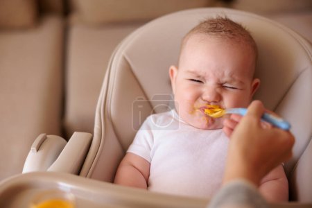 Little baby boy sitting in high chair making funny faces and refusing to eat pumpkin porridge while mother trying to feed him using spoon