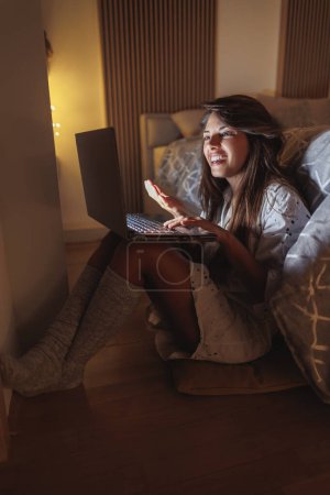 Foto de Hermosa joven sentada en el suelo junto a la cama que tiene videollamada usando computadora portátil mientras se relaja en casa tarde en la noche - Imagen libre de derechos