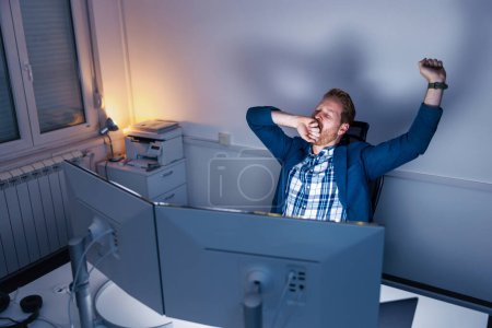 Photo for Tired businessman yawning and stretching at his desk while working late at the office - Royalty Free Image