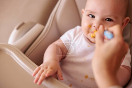 Foto de Madre alimentando al bebé con cuchara, introduciendo la primera comida sólida, el bebé sentado en la silla alta todo desordenado y estabilizado con gachas, sonriendo y comiendo - Imagen libre de derechos