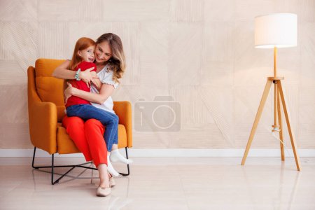 Téléchargez les photos : Mère et fille assises dans un fauteuil, mère tenant son bel enfant sur ses genoux, l'embrassant et la câlinant, appréciant passer du temps libre ensemble à la maison - en image libre de droit