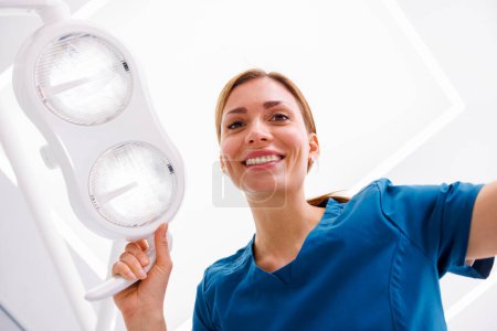 Téléchargez les photos : Vue à angle bas des réflecteurs de réglage des femmes dentistes au-dessus de la tête du patient assis dans une chaise dentaire - en image libre de droit