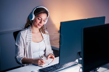 Photo for Business woman sitting at her desk wearing headset having online briefing while working late in an office - Royalty Free Image