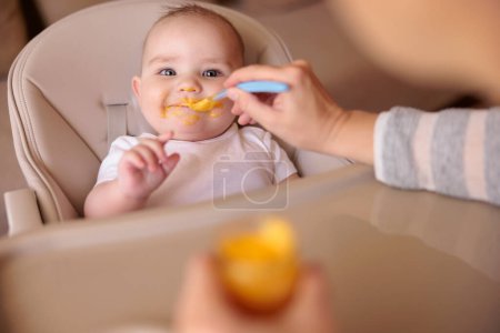 Photo for Mother feeding her cheerful baby boy using spoon, introducing first solid food meal, baby sitting in high chair all messy and staied with porridge, smiling and eating - Royalty Free Image
