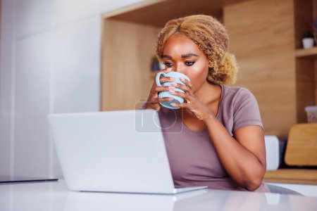 Photo for Woman sitting at kitchen counter drinking coffee and using laptop computer while working remotely from home - Royalty Free Image