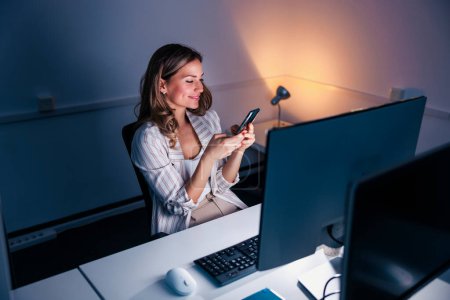 Photo for Casual businesswoman typing text message using smart phone while working overtime late at night in an office - Royalty Free Image