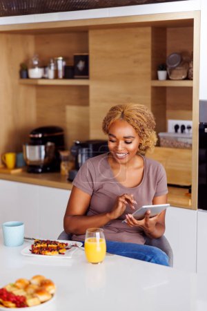 Photo for Young woman sitting at kitchen counter, using tablet computer while having breakfast at home in the morning - Royalty Free Image