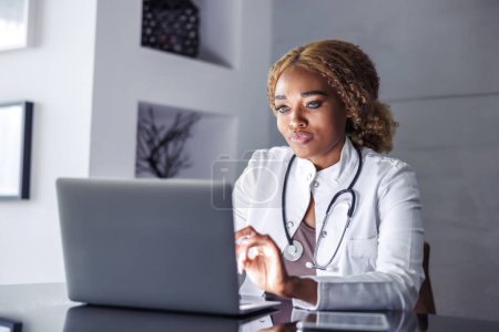 Téléchargez les photos : Médecin femme assise à son bureau à la maison, dactylographiant un rapport médical à l'aide d'un ordinateur portable tout en ayant une consultation par appel vidéo en ligne avec le patient - en image libre de droit