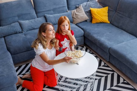 Téléchargez les photos : Vue en angle élevé de la mère et de la fille gaies s'amusant passer du temps libre ensemble à la maison, regarder des dessins animés à la télévision et manger du pop-corn - en image libre de droit
