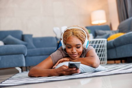 Foto de Mujer acostada en el suelo con auriculares navegando por la red usando un teléfono inteligente mientras se relaja en casa, disfrutando del tiempo libre - Imagen libre de derechos