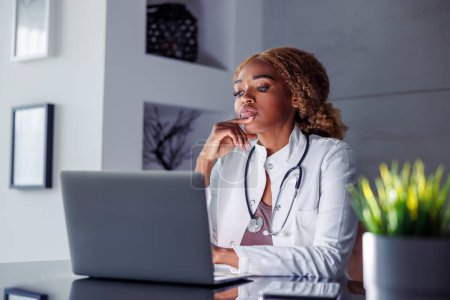 Téléchargez les photos : Médecin femme assise à son bureau à la maison, dactylographiant un rapport médical à l'aide d'un ordinateur portable tout en ayant une consultation par appel vidéo en ligne avec le patient - en image libre de droit