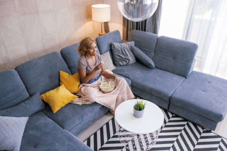 Téléchargez les photos : Vue en angle élevé de belle femme africaine assise sur le canapé du salon, mangeant du pop-corn et regardant un film comique à la télévision, se détendre et profiter du temps libre à la maison - en image libre de droit