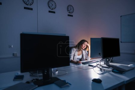 Téléchargez les photos : Fatiguée et stressée femme travaillant tard dans un bureau, penchée à son bureau et tenant la tête dans les mains tout en regardant l'écran d'ordinateur de bureau - en image libre de droit
