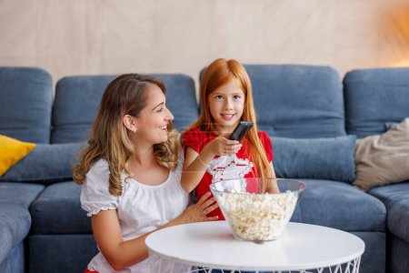 Téléchargez les photos : Mère et fille joyeuses profiter du temps libre ensemble à la maison, regarder la télévision et manger du pop-corn - en image libre de droit