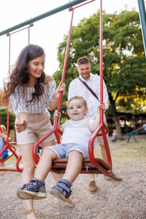 Téléchargez les photos : Joyeux jeune père et mère qui s'amusent à jouer avec leur enfant sur une aire de jeux dans le parc, le balançant sur une balançoire - en image libre de droit