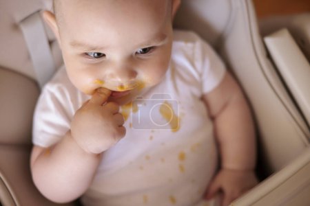 Téléchargez les photos : Vue grand angle de mignon petit garçon sucer doigt après avoir mangé du porridge dans la chaise haute, regardant vers la caméra et souriant - en image libre de droit
