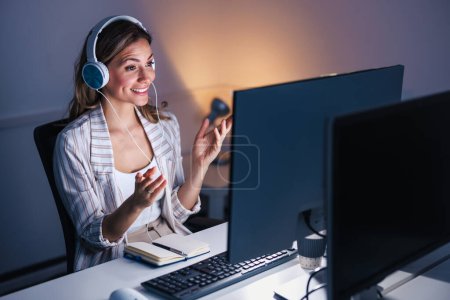 Téléchargez les photos : Femme d'affaires assis à son bureau portant casque ayant appel vidéo à l'aide d'un ordinateur de bureau tout en travaillant des heures supplémentaires dans un bureau - en image libre de droit