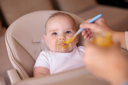 Foto de Lindo bebé niño sentado en silla alta siendo alimentado con gachas por su madre - Imagen libre de derechos