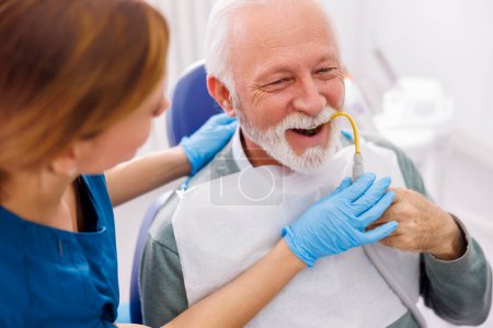 Photo for Senior man sitting at dentist chair holding saliva suction tube while doctor fixing his tooth - Royalty Free Image