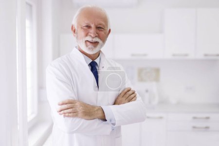 Photo for Portrait of senior male doctor wearing uniform standing in his office in hospital with arms crossed; general practitioner in doctor's office - Royalty Free Image