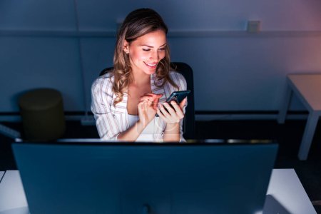 Photo for Casual businesswoman chatting online using smart phone while working overtime late at night in an office - Royalty Free Image
