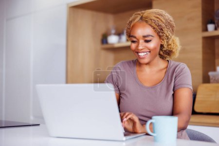 Photo for Woman sitting at kitchen counter drinking coffee and using laptop computer while working from home - Royalty Free Image