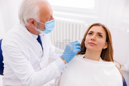 Frau mit Anti-Age-Schönheitsbehandlung in der Arztpraxis; Arzt appliziert Hautfüllstoffe auf weibliches Patientengesicht, korrigiert Falten